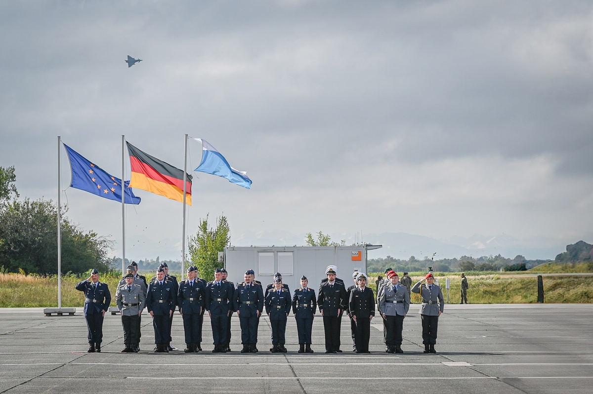 Auf der großen Freifläche stehen die Beförderten. Im Hintergrund wehen die europäische, die deutsche und die Bayern-Flagge im Wind. Ganz hinen sieht man die Alpen. Über der Gruppe fliegt ein Eurofighter durch den wolkenverhangenen Himmel.