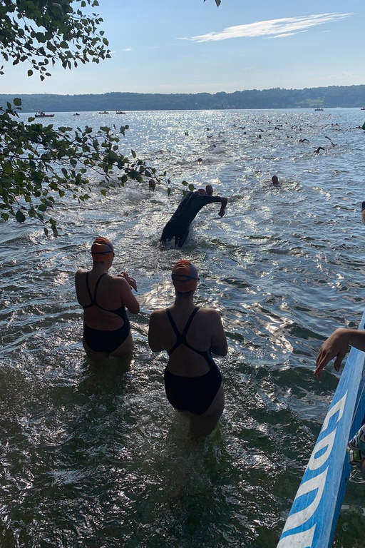 Foto am Seeufer. Zwei Schwimmerinnen stehen im flachen Wasser am Ufer, während ein kleines Stück weiter draußen ein Schwimmer gerade mit den vor sich ausgestreckten Armen ins Wasser springt