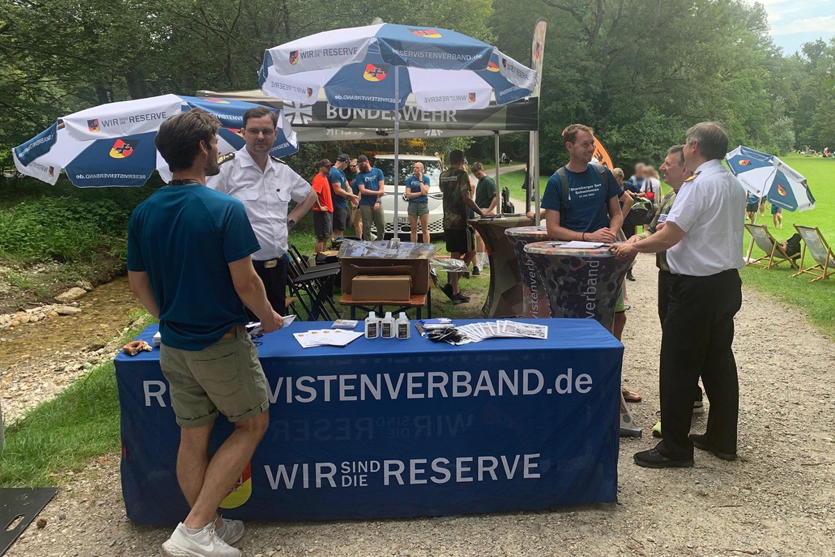 Ein Stand mit Sonnenschirmen, an dem mehrere Menschen stehen und miteinander reden. Vorne an der Stoffverkleidung des Tisches ist der Schriftzug "reservistenverband.de – Wir sind die Reserve" zu lesen.