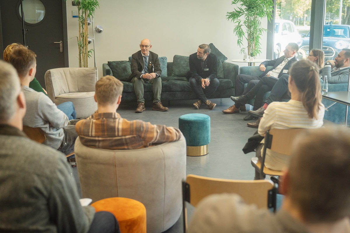 Blick auf eine Sofaecke zwischen dem Publikum hindurchfotografiert. Auf den Sofas sitzen Personen verschiedenen Alters, die Männer tragen Anzüge. Einer spricht gerade zum Publikum. Das Publikum scheint aus jüngeren Menschen zu bestehen.